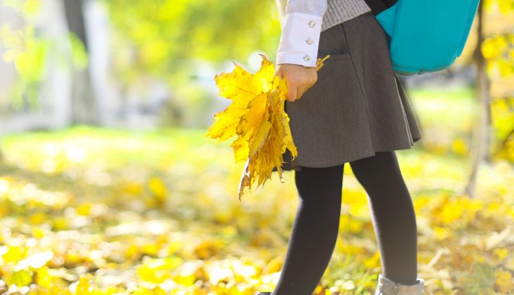 Back to school. Little schooler girl wearing school uniform and stylish blue school backpack having fun with yellow maple leaves in the autumn park